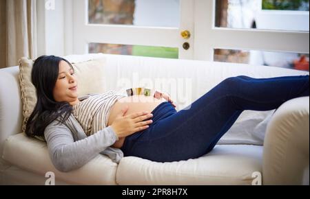 Le début de quelque chose de beau. Une jeune femme enceinte attrayante équilibrant des blocs de bois sur son ventre tout en se relaxant sur le canapé à la maison. Banque D'Images