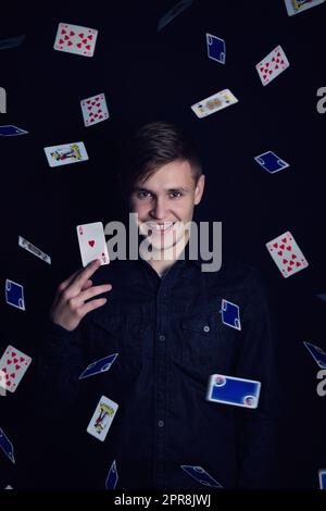 Un jeune homme couronné de succès tient une carte de chance de victoire. Joueur jouer à des jeux de chance et gagner, isolé sur fond sombre. Gagnant de jackpot de jeu avec un as de coeur sous une pluie de cartes de jeu de coeur sous une pluie de cartes de jeu Banque D'Images