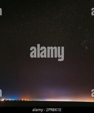 Ciel étoilé de nuit avec étoiles brillantes au-dessus du paysage de la ville de campagne. Rural Field Meadow au début du printemps. Éclat éclatant des étoiles. Illumination Lumières de petite ville ou village. Galaxie Milky Way Banque D'Images