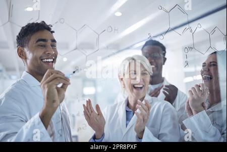 Un groupe diversifié de scientifiques heureux se claquant les mains tout en écrivant et en planifiant ensemble sur un conseil d'administration au travail. Une équipe de professionnels de laboratoire joyeux qui partagent des idées et des brainstorming tout en se tenant ensemble Banque D'Images