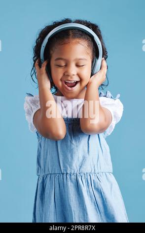 Adorable petite fille hispanique debout avec les yeux fermés et en train de regarder heureux tout en portant un casque sans fil et en appréciant sa chanson préférée sur un fond bleu studio Banque D'Images