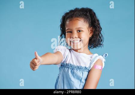 Joyeux enfant vacciné montrant des pouces avec du plâtre sur le bras après l'injection du vaccin debout sur un fond bleu studio. Publicité de vaccination contre le coronavirus. Immunisation des enfants Banque D'Images