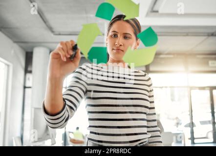 Jeune femme d'affaires spécialisée spécialisée dans les courses mixtes et concentrée qui dessine un symbole de recyclage sur une fenêtre en verre dans un bureau au travail. Un homme d'affaires hispanique qui dessine un signe de sensibilisation au recyclage Banque D'Images