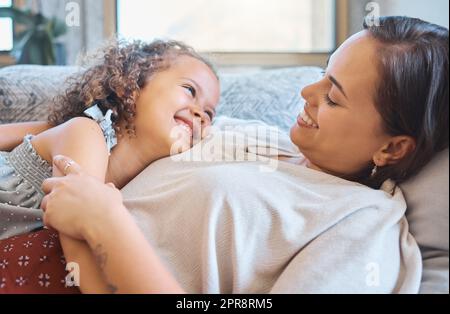 Une petite fille joyeuse regardant le visage de ses mères alors qu'elle latait la tête sur sa poitrine pendant qu'ils se détendent et passent du temps ensemble sur le canapé à la maison en partageant un moment d'appel d'offres Banque D'Images