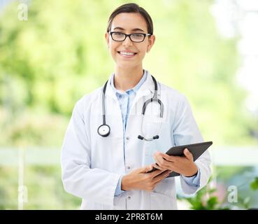 Jeune femme médecin hispanique portant un manteau et souriant tout en utilisant une tablette numérique dans son bureau. La santé physique est importante pour votre bien-être. Médecin de race mixte utilisant un appareil sans fil à son travail à l'hôpital Banque D'Images