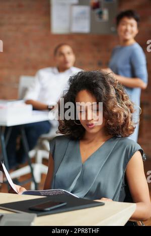 En lisant la dernière proposition commerciale, une jeune femme d'affaires passe par la paperasse dans un bureau avec ses collègues en arrière-plan. Banque D'Images