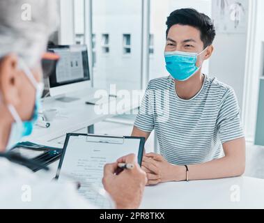 Remplir le formulaire obligatoire. Un jeune homme portant un masque et assis avec son médecin pendant sa consultation à la clinique. Banque D'Images