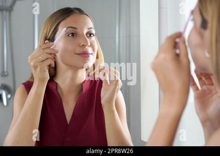 Gros plan sur une femme latine rasant ses sourcils avec un rasoir dans le miroir à la maison Banque D'Images