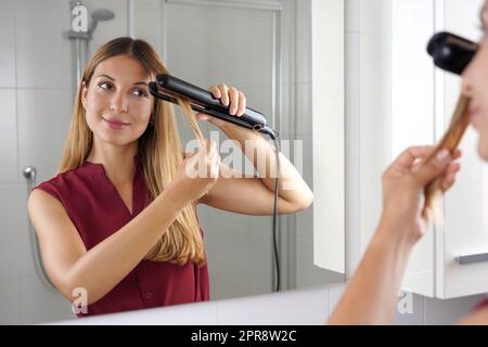 Fille utilisant un lisseur vapeur pour coiffer les cheveux au miroir de la salle de bains Banque D'Images