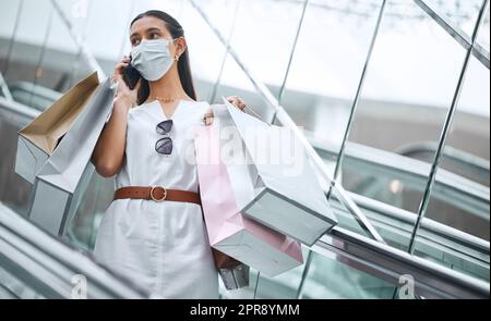 Une jeune femme mixte de race portant un masque médical et parlant sur un téléphone portable alors que sur un escalator après une frénésie de shopping. hispanique à la mode portant des sacs de détail après avoir acheté dans un centre commercial pendant la pandémie Covid-19 Banque D'Images