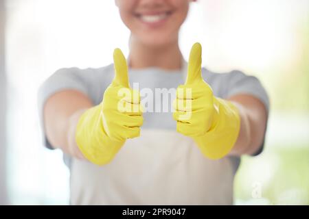 Un travailleur domestique méconnaissable montrant les pouces tout en portant des gants et en nettoyant. Une femme de race mixte inconnue qui a l'air heureuse lors du nettoyage de son appartement Banque D'Images