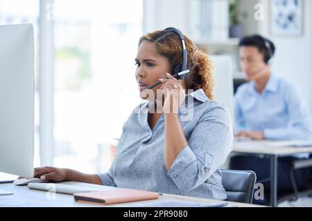 Un agent de télémarketing de centre d'appels de course mixte stressé et confus qui parle sur un micro-casque tout en travaillant sur un ordinateur dans un bureau. Jeune femme afro-américaine choquée traitant de crise de panique et appelant difficile pour l'assistance à la clientèle Banque D'Images