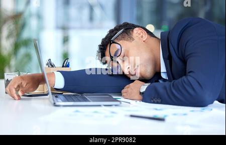 Un homme d'affaires de course mixte épuisé portant des lunettes tout en dormant à son bureau. Jeune entrepreneur se sentant surtravaillé, fatigué et démotivé. Un homme paresseux se lapiait et ignorait les délais en faisant une sieste. Épuisement professionnel et stress sur le lieu de travail Banque D'Images