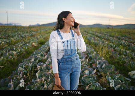 Femme agriculteur parlant sur son smartphone en se tenant dans un champ de chou. Jeune femme brune utilisant son appareil mobile sur une ferme végétale biologique Banque D'Images