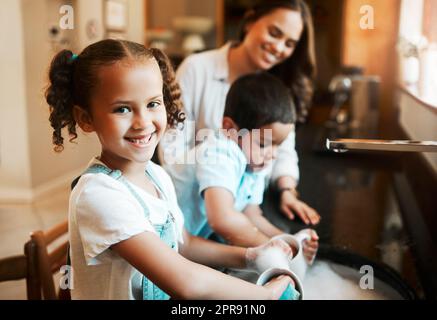 Bonne joyeuse petite fille de race mixte aidant sa mère à laver les plats dans la cuisine à la maison. Enfant hispanique souriant tout en lavant une tasse avec du savon et de l'eau. La famille garde leur maison propre Banque D'Images