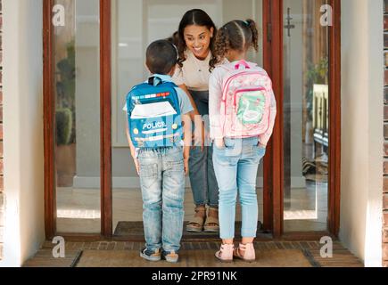 Jeune mère voyant ses enfants à l'école. Race mixte disant au revoir à son fils et à sa fille alors qu'ils quittent la maison pour l'école. Bonne maman saluant ses enfants à la porte d'entrée de leur maison Banque D'Images