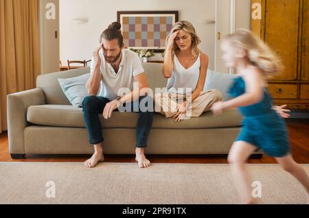 La jeune mère et le père caucasiens souffrent d'un mal de tête avec leurs filles qui s'exécutent rapidement dans le salon à la maison. Petits frères et sœurs jouant ensemble tandis que leurs parents sont fatigués. Femme et homme se sont contrariés assis sur le canapé Banque D'Images