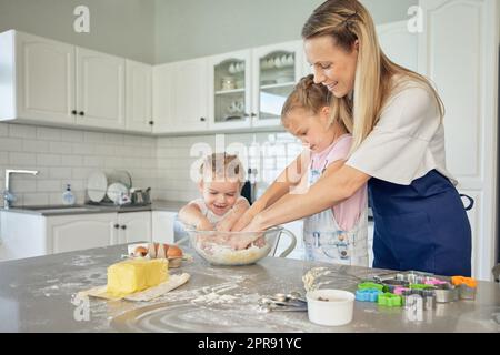 La mère et les petites filles caucasiennes cuisent ensemble dans une cuisine à la maison. Maman enseignant aux filles comment faire de la pâte dans une cuisine désordonnée. Les sœurs apprennent à cuire avec leur mère Banque D'Images