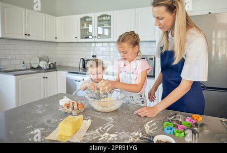 Mère caucasienne et petites filles cuisant ensemble dans une cuisine à la maison. Maman enseignant aux filles comment faire de la pâte dans une cuisine désordonnée. Les sœurs apprennent à cuire avec leur mère Banque D'Images