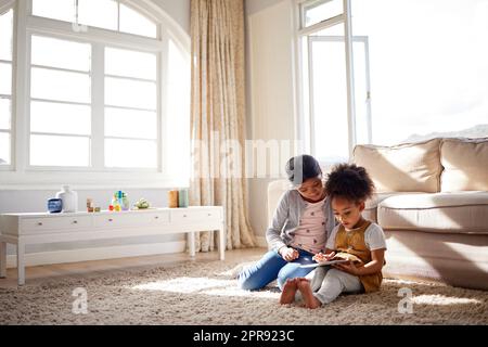 Deux petites sœurs de race mignonne assis sur le tapis et jouant ensemble sur une tablette numérique à la maison. Adorables amies hispaniques qui apprennent à utiliser un appareil sans fil ensemble Banque D'Images