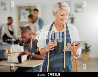 La designer de mode senior lit les commandes en ligne d'une tablette portable dans sa boutique. Une vieille femme productrice de vêtements souriant de son succès tout en tenant le café. Un entrepreneur créateur mûr heureux Banque D'Images