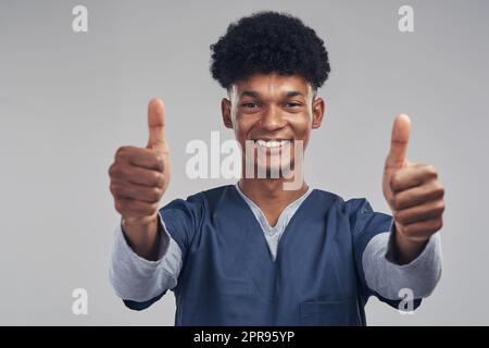 Je suis ici pour vous donner de bonnes nouvelles. Une infirmière masculine montrant les pouces vers le haut tout en se tenant debout contre un fond gris. Banque D'Images