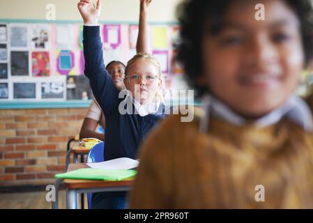 Je connais la réponse. Un groupe diversifié d'enfants assis dans leur salle de classe et levant les mains pour répondre à une question. Banque D'Images