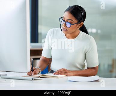 Y a-t-il autre chose dont vous pourriez avoir besoin : une jeune femme d'affaires attirante assise seule dans son bureau et écrivant des notes tout en portant un casque ? Banque D'Images