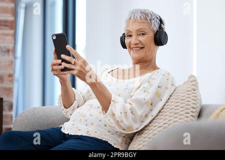 Le petit appareil de divertissement le plus petit. Une femme âgée utilisant un smartphone et un casque sur le canapé à la maison. Banque D'Images