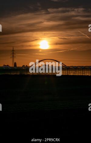 Coucher de soleil doré sur les silhouettes de serre avec pont et tour d'électricité pour l'énergie solaire dans les affaires agricoles sur la campagne idyllique et le paysage rural montre serres de verre des légumes sains Banque D'Images