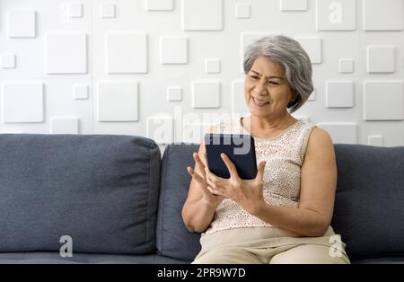 Asiatique cheveux gris vieille femme regardant la tablette écran d'ordinateur, souriant tout en s'asseyant sur le canapé dans le salon. Banque D'Images