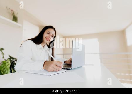 Femme assise au bureau, lisant depuis un ordinateur portable, écrivant des notes sur son ordinateur portable Banque D'Images