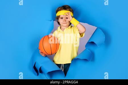 Un enfant souriant avec un ballon de basket-ball montrant un signe ok, un excellent symbole. Enfant garçon avec boule de panier regardant à travers trou de papier. Jeu de basket-ball. Petit Banque D'Images