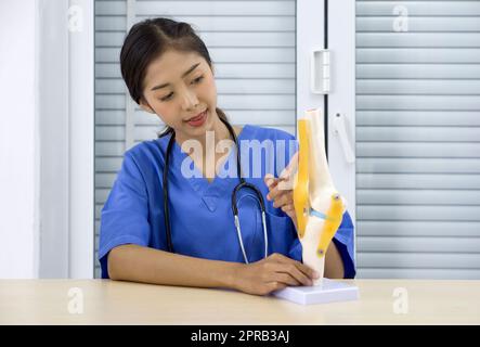 Jeune médecin asiatique en uniforme bleu et stéthoscope pointant vers le ligament croisé antérieur sur le modèle d'articulation fonctionnelle de genou de luxe. Banque D'Images