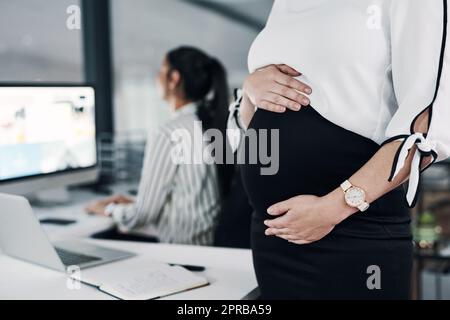 La maternité ne m'empêche pas d'atteindre des objectifs. Une femme d'affaires enceinte méconnue se tenant debout et tenant son estomac pendant qu'une collègue travaille derrière elle. Banque D'Images