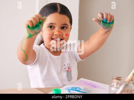 Plus elle devient messieuse, plus elle est amusante. Une adorable petite fille assise à la maison avec de la peinture à base d'eau enduite ses mains. Banque D'Images