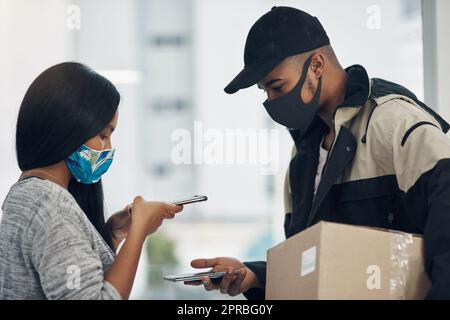 Le paiement sans contact permet de garder le contrôle. Un jeune homme et une jeune femme masqués utilisant un smartphone lors d'une livraison à domicile. Banque D'Images