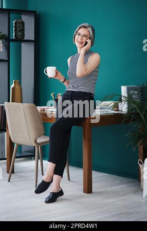 Il me fait sentir bien d'entendre les clients. Une femme d'affaires ayant le café tout en parlant sur son téléphone portable dans son bureau. Banque D'Images