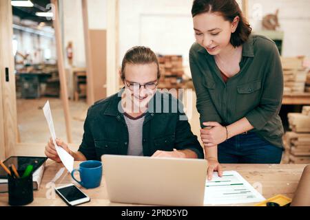 Tout va parfaitement selon le plan. Deux jeunes charpentiers planifiant et travaillant ensemble sur un ordinateur portable dans leur atelier. Banque D'Images