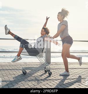 Les bons amis sont le plus grand cadeau. Une jeune femme poussant son ami autour de la promenade dans une carte de shopping. Banque D'Images