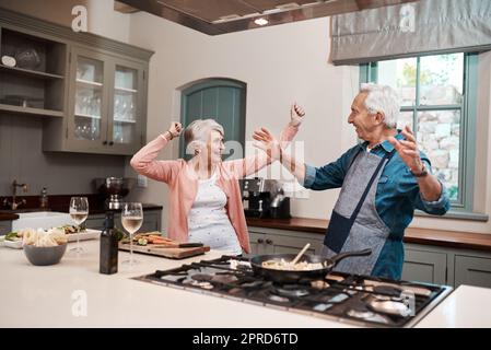 Une bonne cuisine met tout le monde dans une bonne humeur. Un couple de personnes âgées dansant tout en cuisinant dans la cuisine à la maison. Banque D'Images