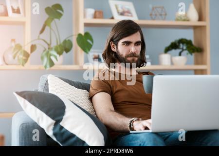 En regardant de plus près les choses, un beau jeune homme utilisant son ordinateur portable tout en se relaxant sur un canapé dans son salon. Banque D'Images