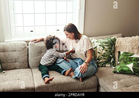 Nous avons un peu de médecin en fabrication. une jeune femme gaie et son fils jouant autour avec un stéthoscope tout en étant assis sur un canapé à la maison pendant la journée. Banque D'Images