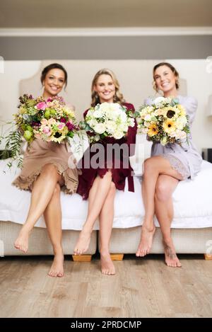 Les jours seront aussi beaux que ces fleurs. Portrait complet de trois jeunes demoiselles d'honneur tenant des bouquets de fleurs tout en étant assis sur un lit dans leur dressing. Banque D'Images