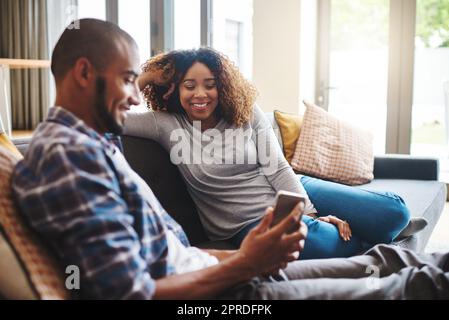 Couple heureux, insouciant et détendu lisant le texte sur un téléphone tout en étant assis sur un canapé. Homme et femme souriant et riant tout en naviguant sur une application de réseaux sociaux en ligne ou en regardant des vidéos amusantes à la maison Banque D'Images