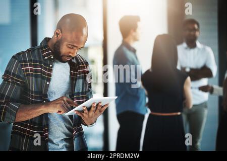 HES travaille toujours intelligemment. Un jeune homme d'affaires utilisant sa tablette numérique dans un bureau moderne avec ses collègues en arrière-plan. Banque D'Images