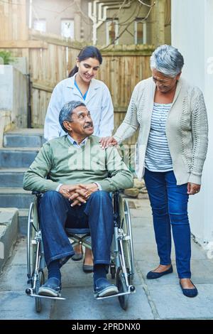 Où que vous alliez, nous allons. Un homme âgé gai assis dans un fauteuil roulant tout en étant soutenu par sa femme et une infirmière féminine à la maison pendant la journée. Banque D'Images