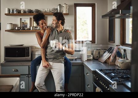 Heureux, dans l'amour et rire tandis qu'un couple interracial aime le café du matin et le collage tout en ayant une bonne communication dans une relation. Mari et femme parlant en se tenant dans la cuisine à la maison Banque D'Images
