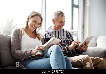 En mode de relaxation du dimanche, une femme mûre lisant un livre tandis que son mari utilise une tablette numérique sur le canapé à la maison. Banque D'Images