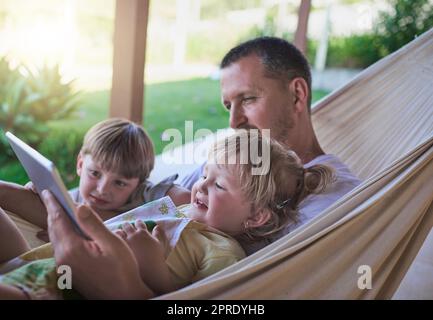 Les pères sont toujours prêts à regarder des dessins animés avec leurs enfants. Un père et ses deux enfants en utilisant une tablette numérique tout en se relaxant sur un hamac en plein air. Banque D'Images